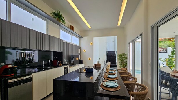 Een moderne keuken met een centraal eiland, zwarte aanstootbladen, en een eethoek met rieten stoelen en een tafel met glazen blad.
