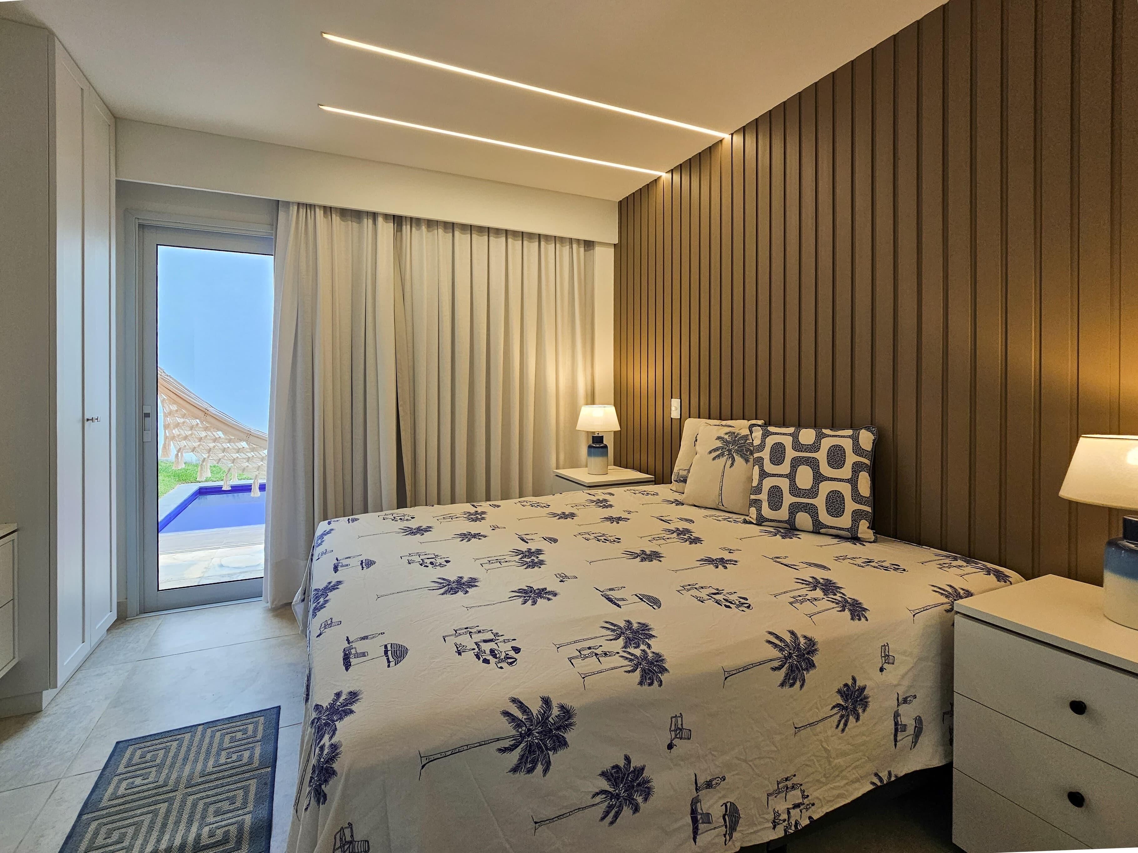 A hotel room with a bed, two bedside tables, a window with curtains, and a view of a pool and building outside.