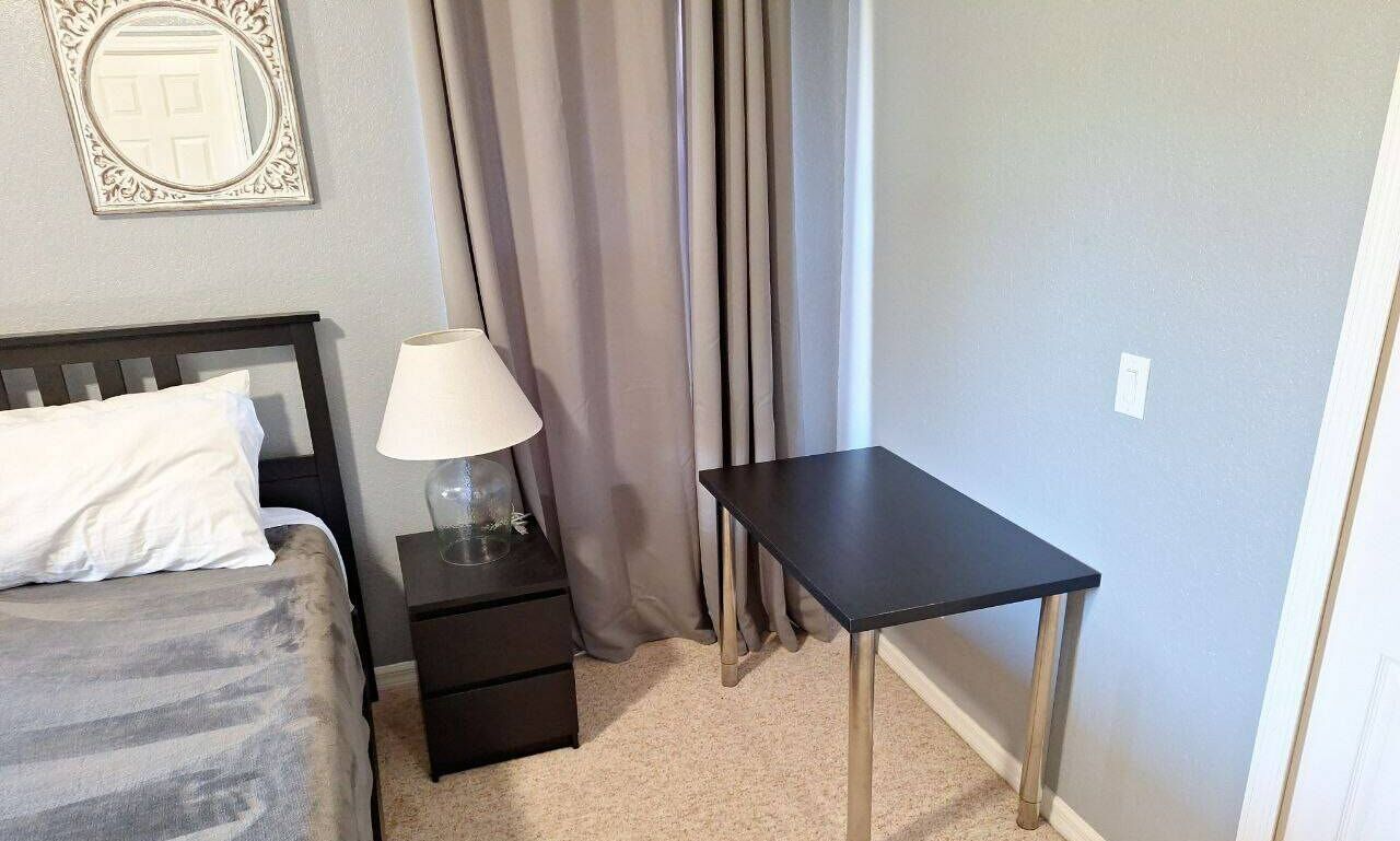 Un dormitorio con una cama, una mesita de noche con lámpara, una pequeña mesa y un espejo en la pared.