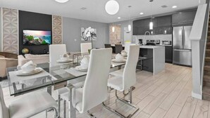 A modern kitchen with a central island, built-in appliances, and a dining area with glass tables and white chairs. - Beautiful Vacation Home With Splash Pool 35 (Kissimmee)