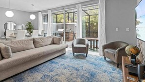 A modern living room with a large sofa, a coffee table, and a TV. There is a large window with curtains, a plant, and a view of the outdoors. - Beautiful Vacation Home With Splash Pool 35 (Kissimmee)