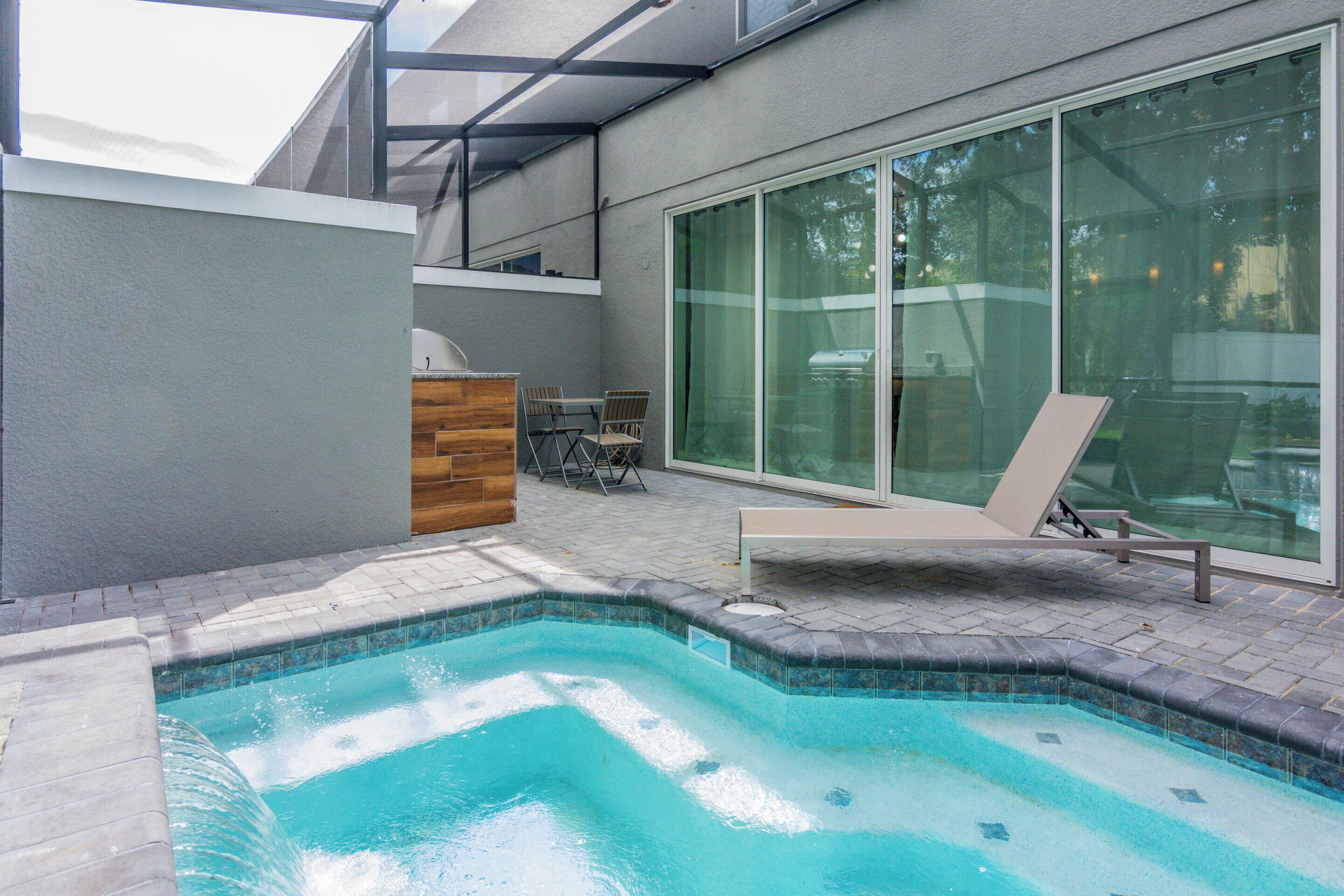 A swimming pool with a wooden deck, a lounge chair, and a glass door leading to a patio area.