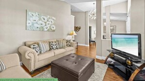 A living room with a beige sofa, a brown ottoman, a flat-screen TV, and a painting on the wall. - Enchanting House few min Away From the Parks 52 (Kissimmee)