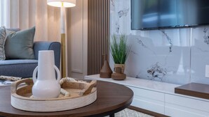 Un salon moderne avec une table basse ronde en bois, un vase blanc et un plateau rempli d’assiettes. Un grand téléviseur à écran plat est fixé au mur, et un canapé avec des coussins se trouve au fond de la pièce.