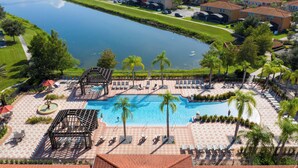 A swimming pool with lounge chairs and palm trees, surrounded by a brick patio and a view of a lake. - Lake View Home With Private Pool at Bella Vida 91 (Kissimmee)