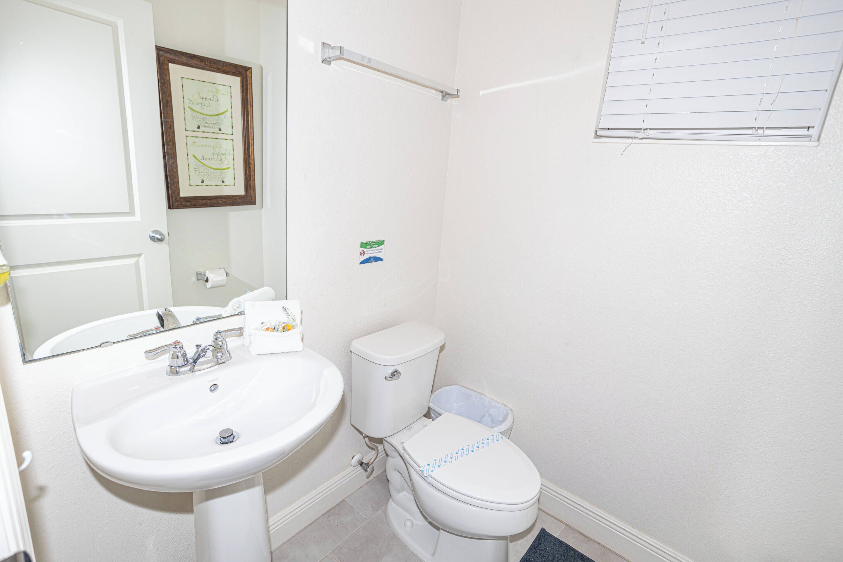 A small, clean bathroom with a white sink, toilet, and mirror. There is a framed picture on the wall and a window with blinds.