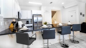 A modern kitchen with white cabinets, a stainless steel refrigerator, and a black countertop. There are four black bar stools and a small island with a sink.