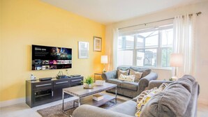 A living room with a flat-screen TV mounted on the wall, a grey sofa set, a coffee table, and a window with curtains.