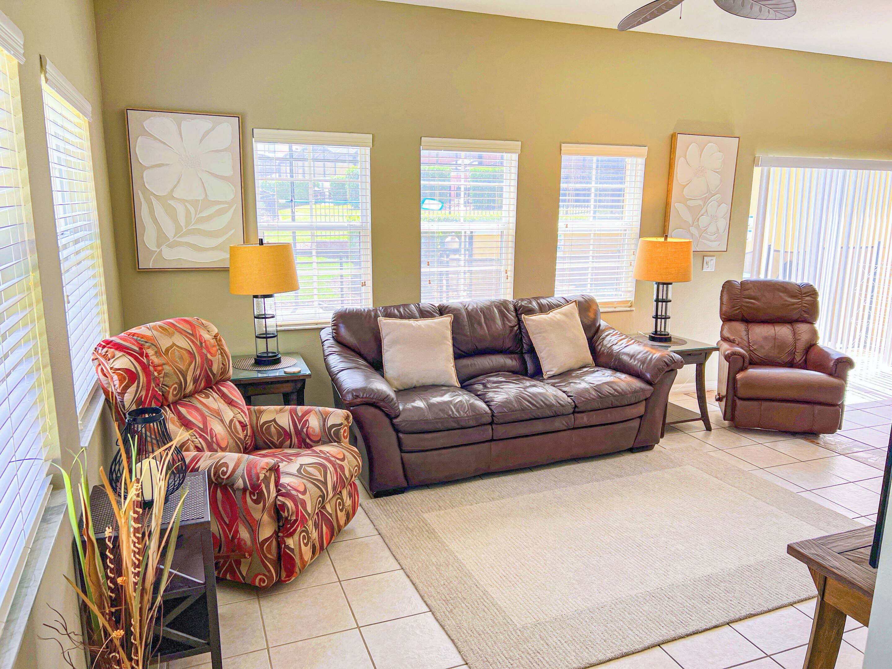 A living room with a brown leather sofa, a patterned armchair, and a recliner. There are two lamps on side tables, and artwork on the walls.