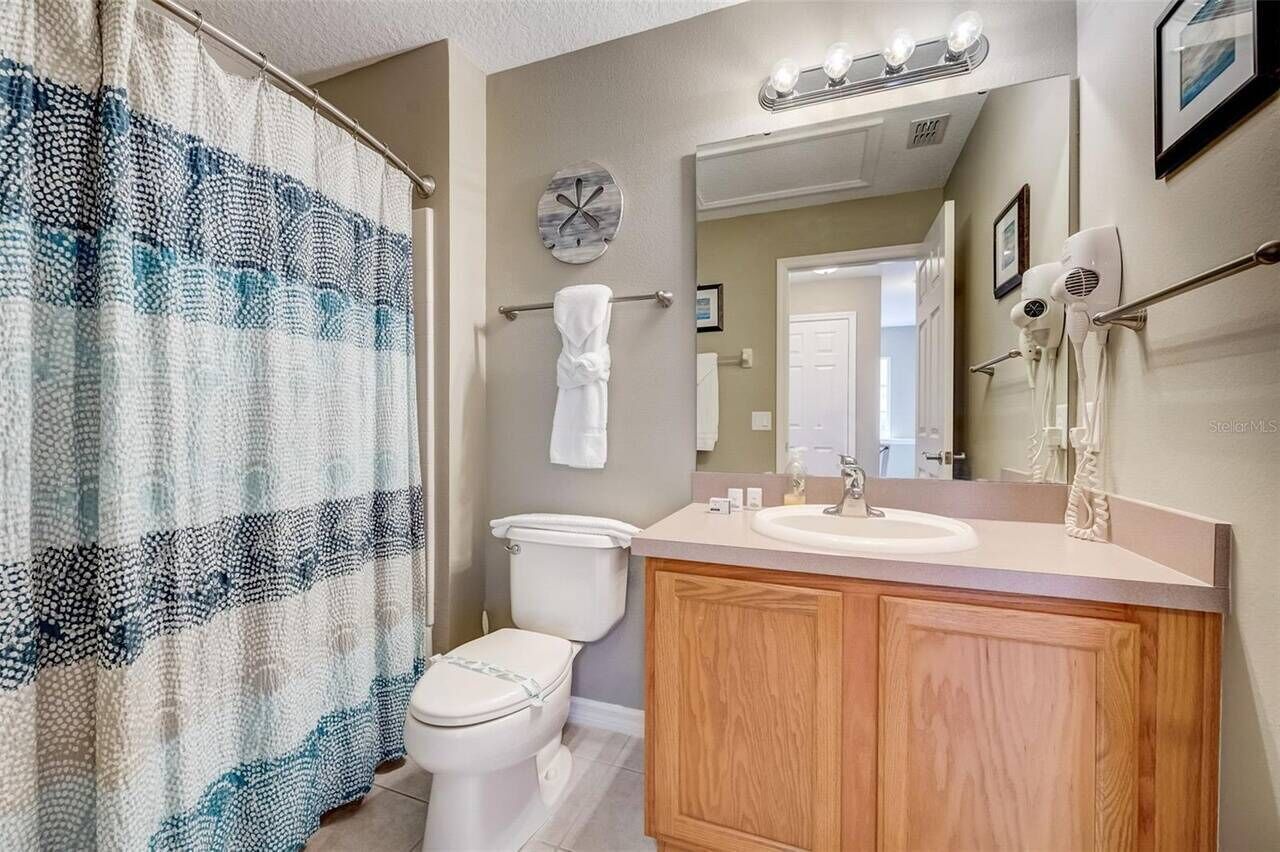 A bathroom with a shower curtain, toilet, vanity with a mirror, and hair dryers on the wall.