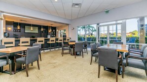 A modern dining area with tables and chairs, a bar area with shelves of bottles, and large windows offering a view of palm trees and buildings.