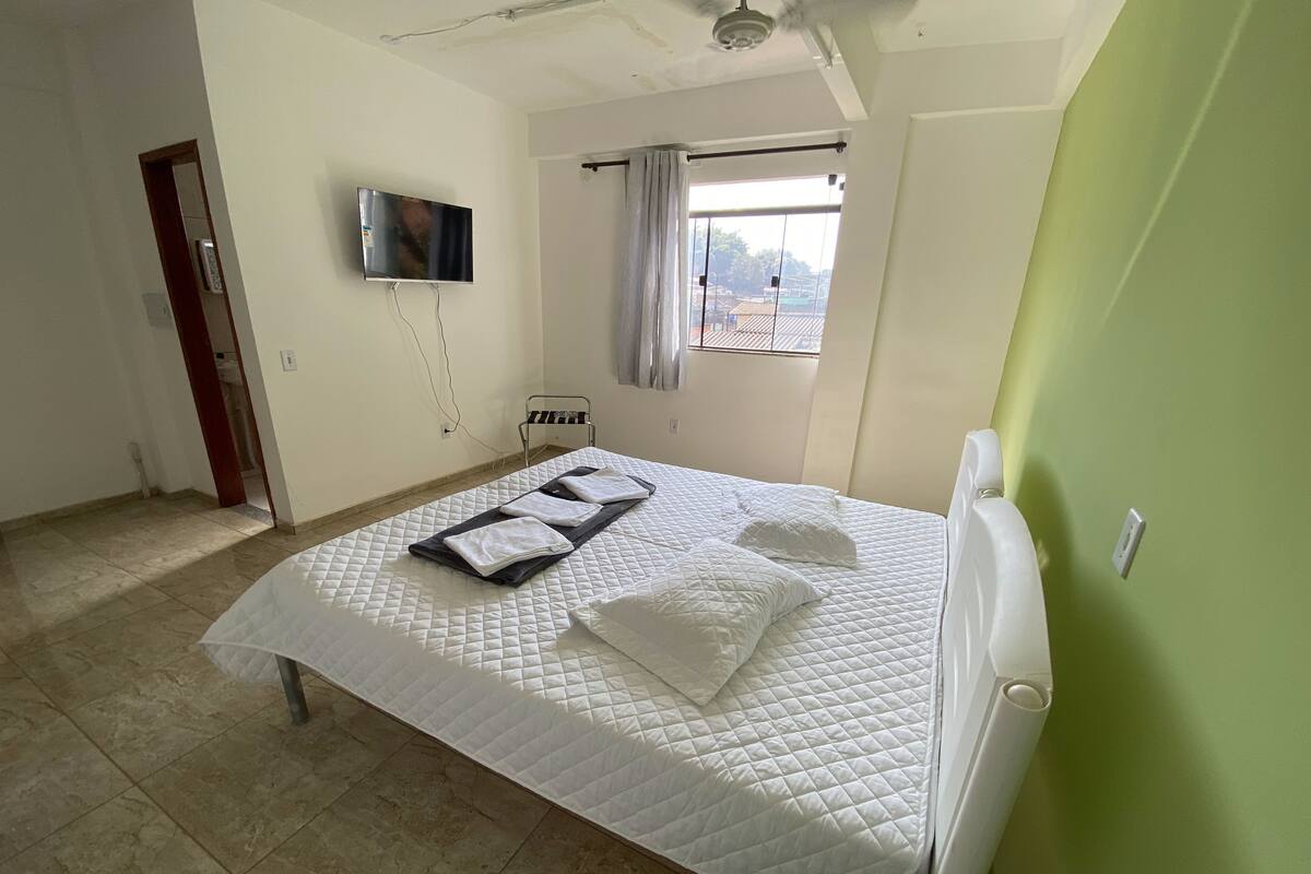 A neatly made bed with pillows and a blanket, a television mounted on the wall, a window with curtains, and a radiator.