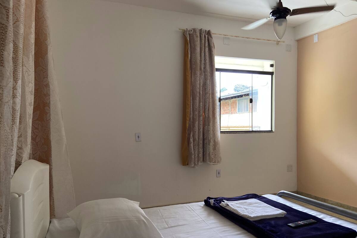 A small, single-bed room with a window, a ceiling fan, and a white pillow on the bed.