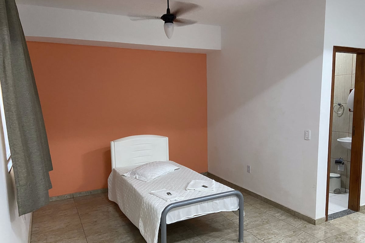 A small, single-bed room with a white headboard, orange walls, and a tiled floor.