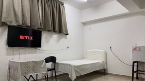 Una habitación pequeña y limpia con una cama, un escritorio con silla, un televisor en la pared y una ventana con cortinas.