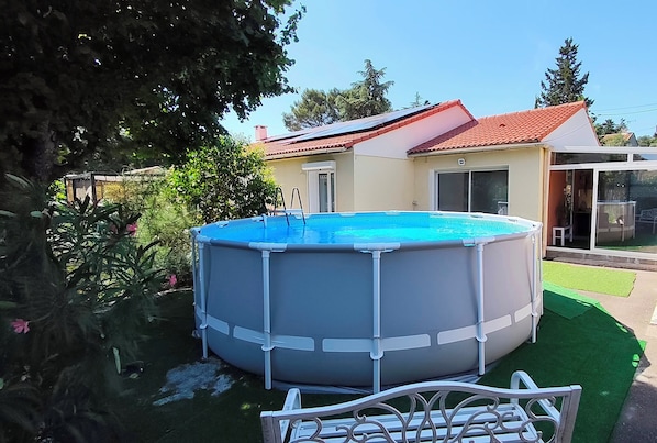 An above-ground swimming pool with a ladder, surrounded by a well-maintained lawn and a house with a red roof.