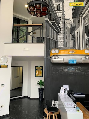 A hotel reception area with a reception desk, a clock, a reception sign, and a wall mural of a street scene. - Hotel Del Medio (Sutomore)