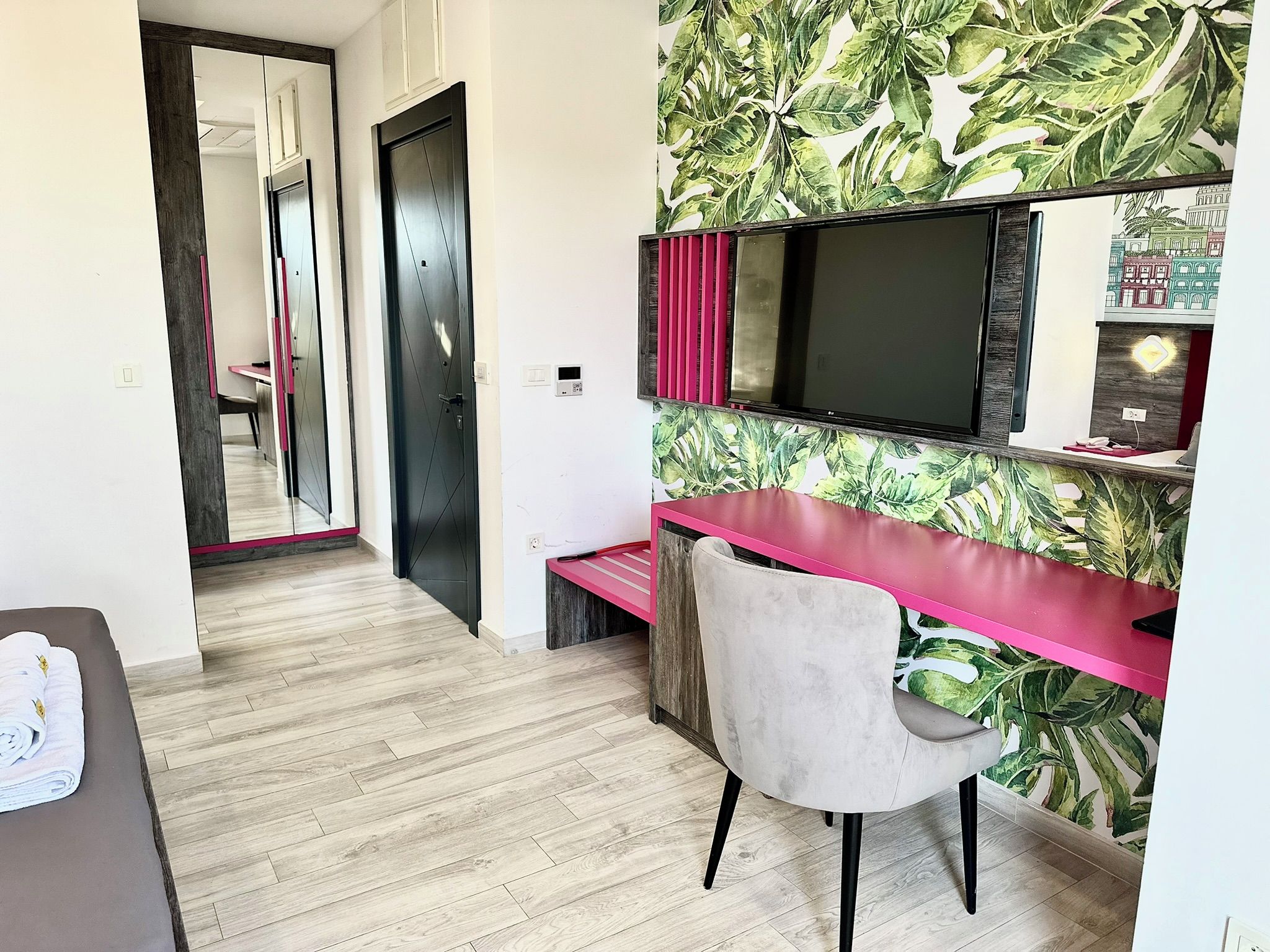 A modern room with a wooden floor, a desk with a pink top, a chair, a large mirror, and a TV.