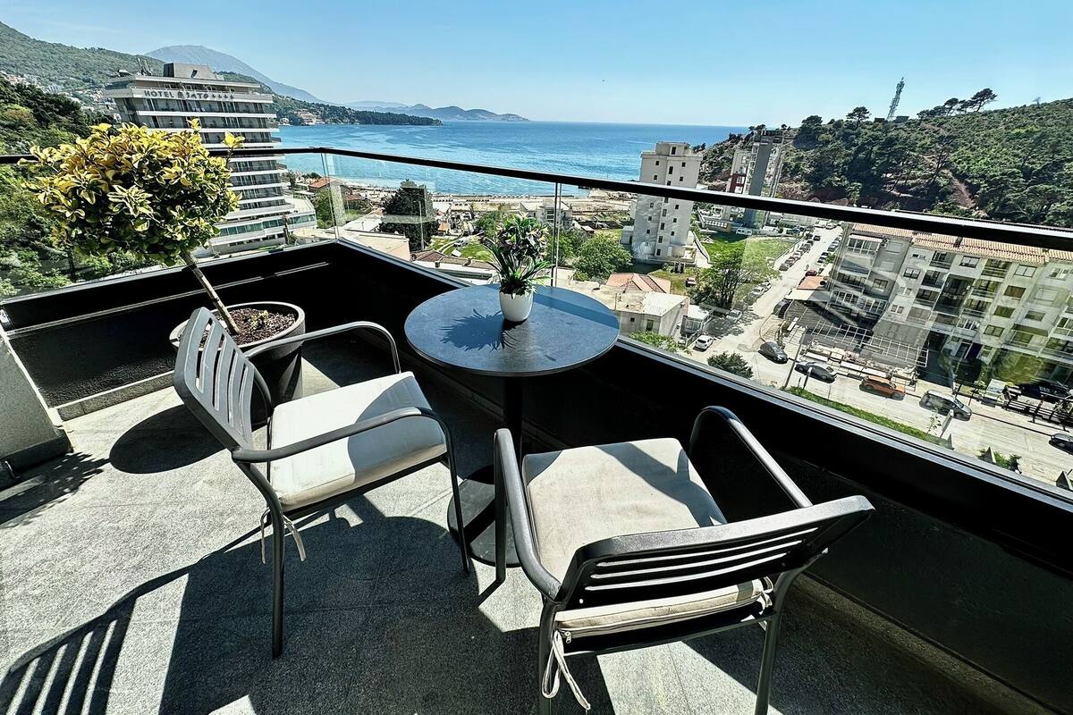 Un balcón con mesa y sillas, con vistas a un paisaje urbano costero con edificios y cielo despejado.