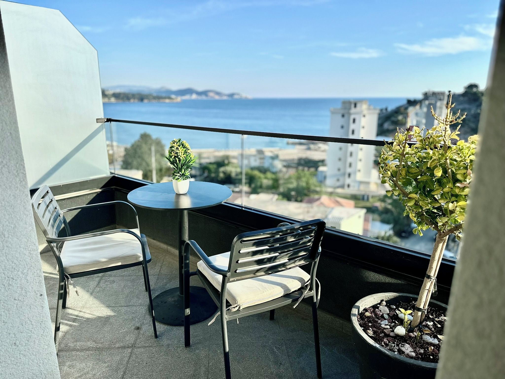 Un balcón con una mesa redonda y dos sillas, una planta en maceta y vistas al mar y edificios.