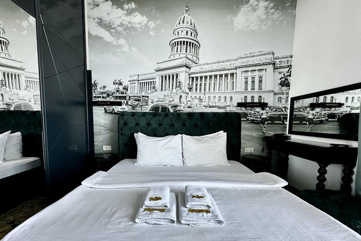 Habitación de hotel con una cama grande, dos almohadas blancas y dos toallas dobladas con logotipos del hotel. Un ventanal muestra un paisaje urbano con un edificio destacado.