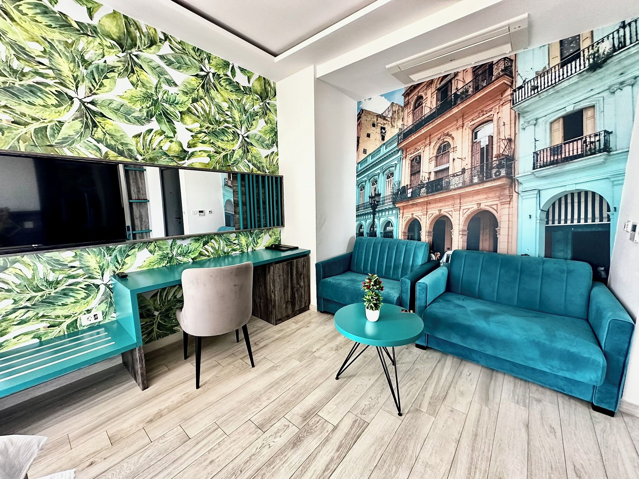 A modern living room with a large flat-screen TV, a teal sofa, a small round table with a plant, and a vibrant leaf-patterned wall.