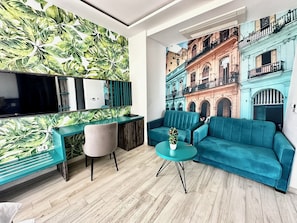 En moderne stue med et stort fladskærms-tv, en turkis sofa, et lille rundt bord med en plante, og en væg med et livligt blad-mønster.
