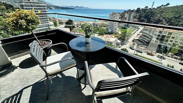 A balcony with a table and chairs, overlooking a coastal cityscape with buildings and a clear sky. - Hotel Del Medio (Sutomore)