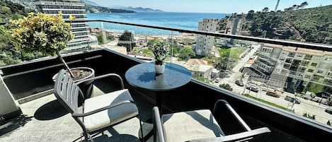 A balcony with a table and chairs, overlooking a coastal cityscape with buildings and a clear sky.