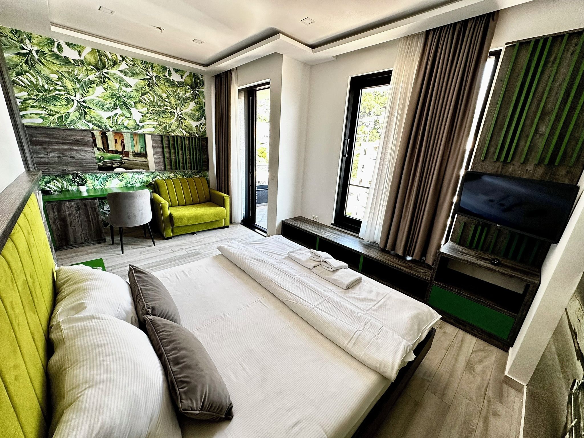 A modern hotel room with a large bed, a desk with a chair, a sofa, and a TV mounted on the wall.