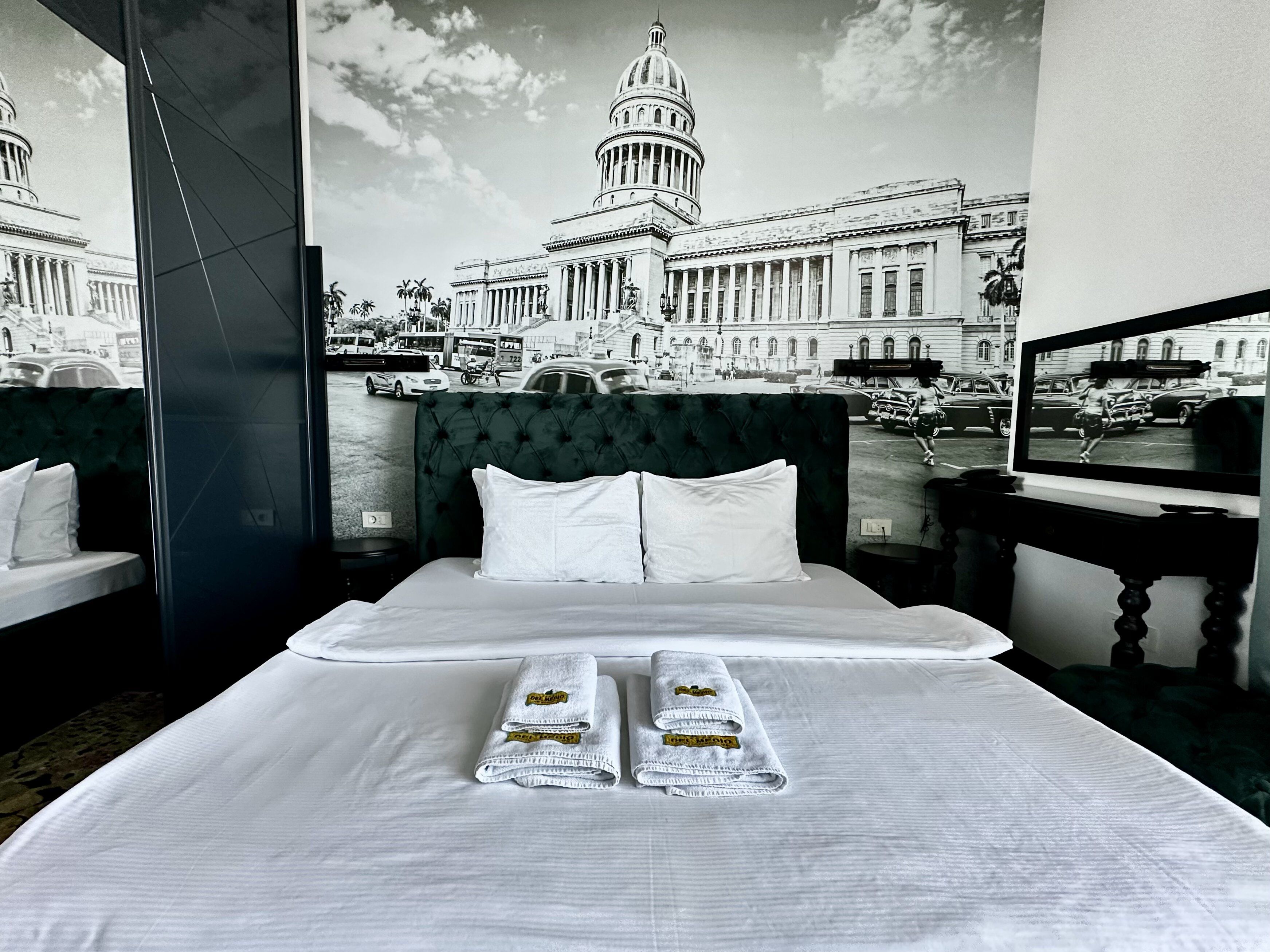 A hotel room with a large bed, two pillows, and two towels with hotel logos. A large window offers a view of a historic building and a street scene.
