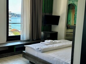 A hotel room with a bed, a TV, a window with curtains, and a view of the sea. - Hotel Del Medio (Sutomore)