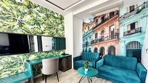 A modern living room with a large flat-screen TV, a teal sofa, a small round table with a plant, and a vibrant leaf-patterned wall. - Hotel Del Medio (Sutomore)
