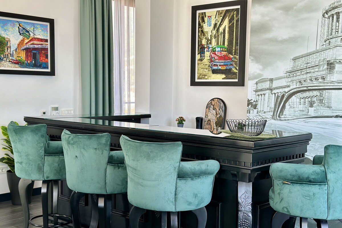 A modern bar area with a long counter, four bar stools, and framed artwork on the walls.