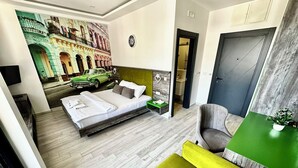 A modern hotel room with a bed, a green sofa, a small table, and a mural of a street scene. - Hotel Del Medio (Sutomore)