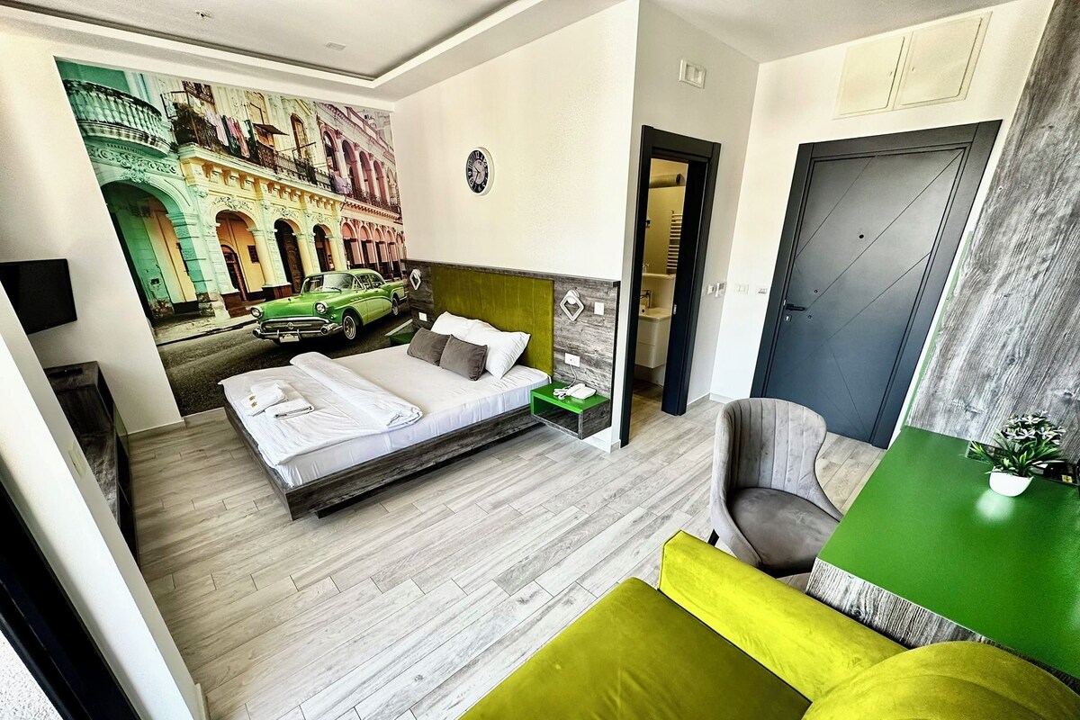 A modern hotel room with a bed, a green sofa, a small table, and a mural of a street scene.