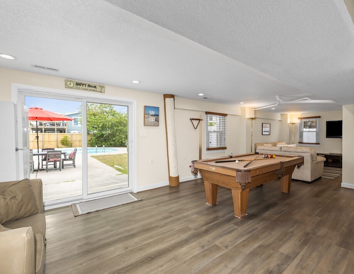 A pool room with a pool table, a sofa, and a sliding glass door leading to an outdoor patio with a pool and furniture.
