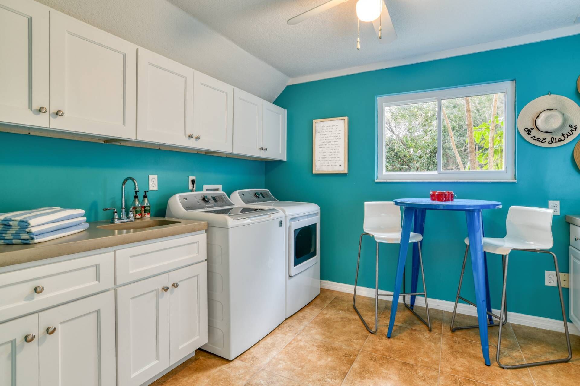A laundry room with a washing machine, a sink, and a window with a view of trees. There are two chairs and a table with a red object on it.