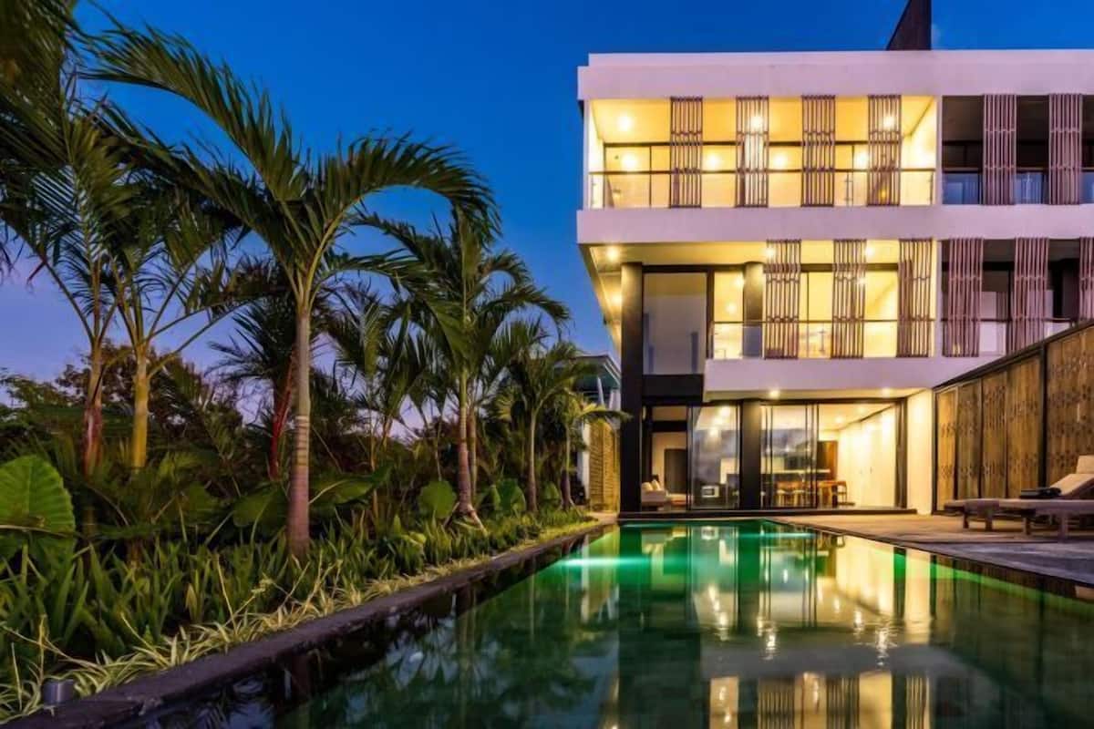 A modern two-story building with a swimming pool, surrounded by tropical vegetation.