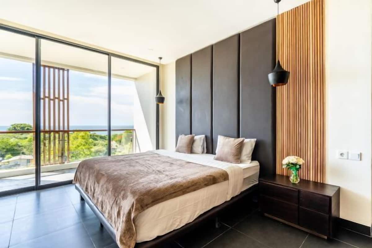 A modern bedroom with a large bed, a nightstand, a wall-mounted lamp, and a view of greenery through a large window.