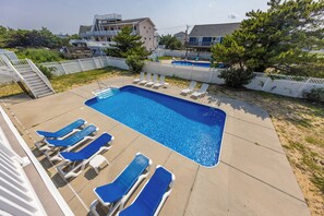 A rectangular swimming pool with a clear blue water surface, surrounded by a concrete deck. There are multiple white lounge chairs with blue cushions arranged around the pool.