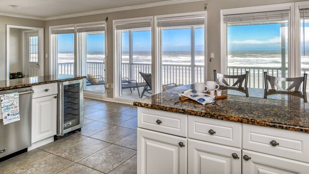 A kitchen with large windows overlooking the beach, a dining area with chairs, and a kitchen island. - High Tide Paradise (St. Augustine)