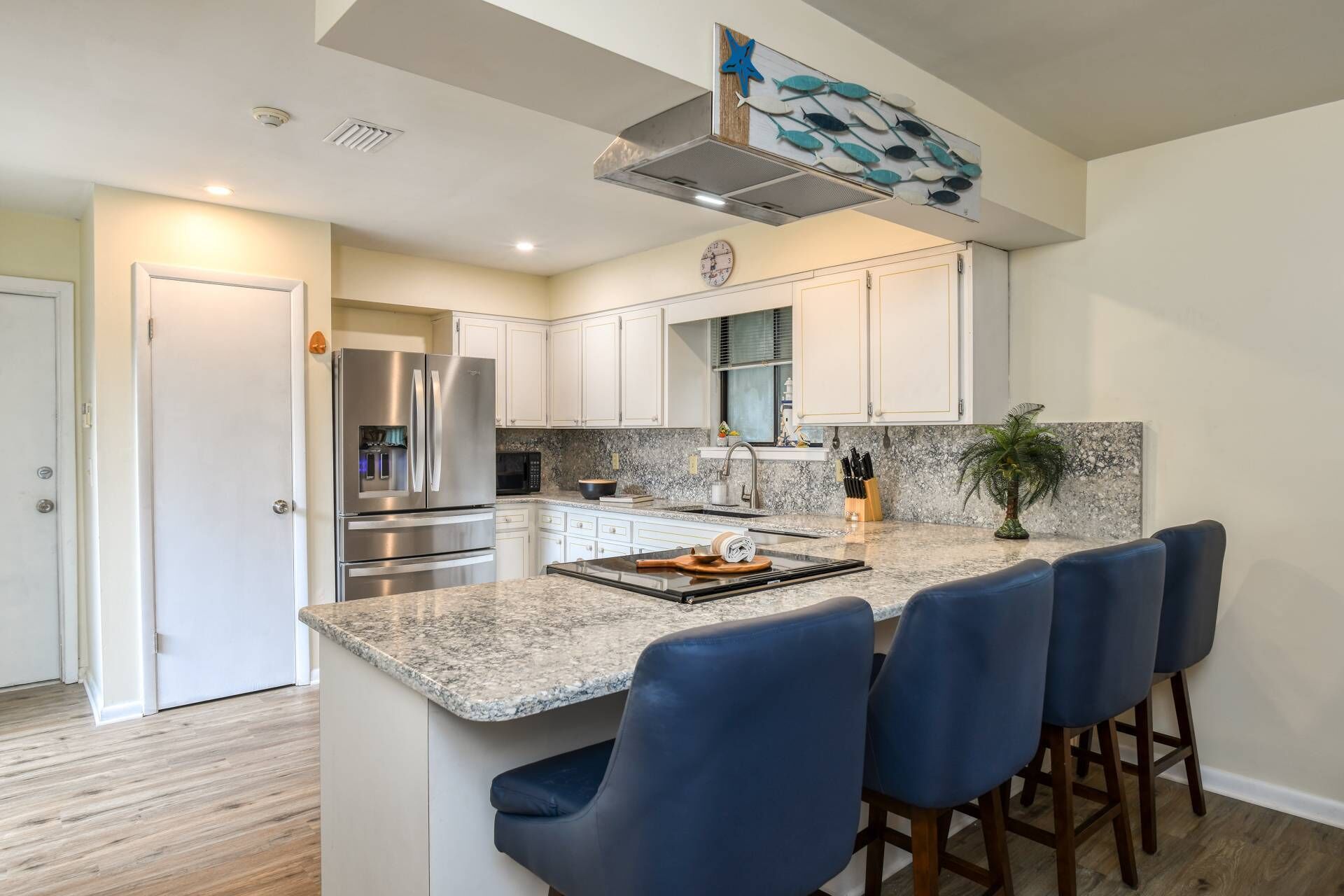 A modern kitchen with white cabinets, a stainless steel refrigerator, a granite countertop, and blue bar stools.