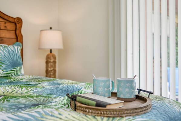A bed with a tropical leaf-patterned bedspread, a wooden headboard, a bedside lamp, and a tray with a book, two star-patterned mugs, and a spoon.