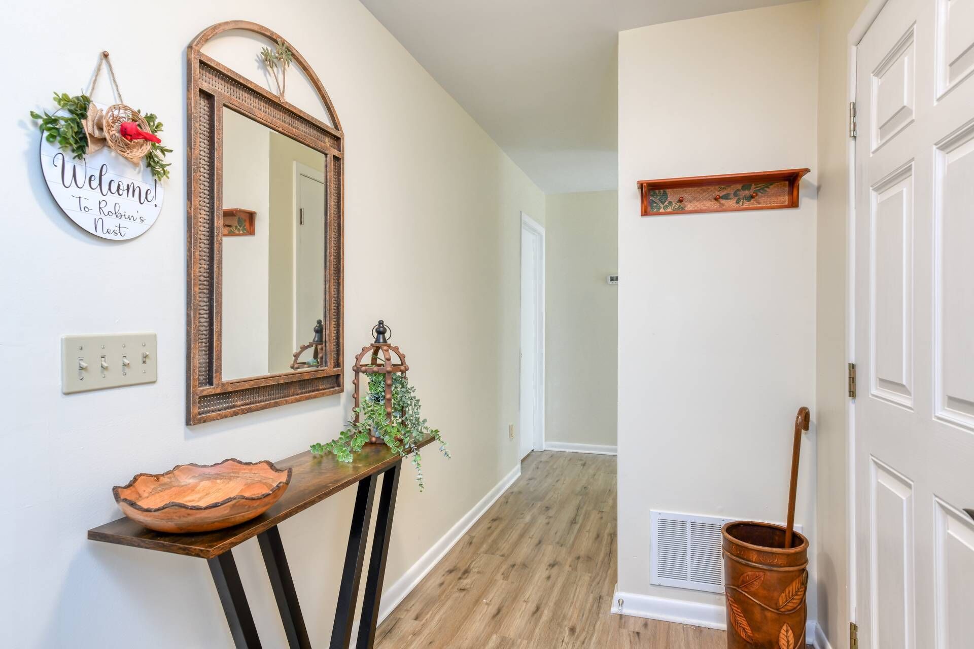 Un couloir avec une console en bois, un miroir, un panneau d’accueil, une petite plante et une porte ornée d’une étagère décorative.