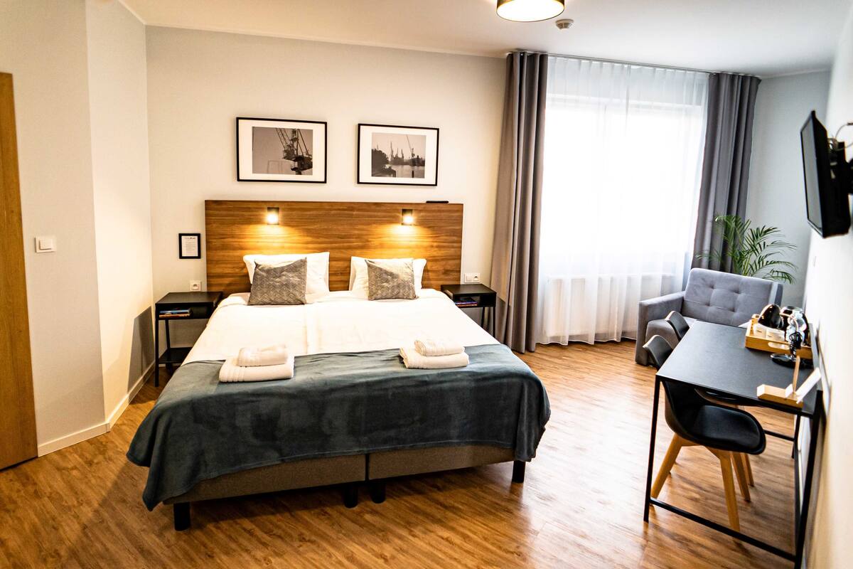 Et hotelværelse med en stor seng, et skrivebord og en stol. Der er to billeder hængende på væggen over sengen.