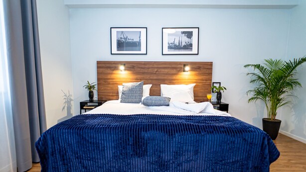 A modern bedroom with a wooden headboard, two framed pictures, a blue bedspread, and a potted plant.