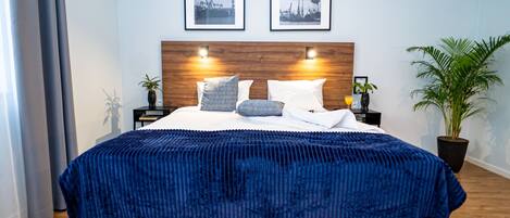A neatly made bed with a blue cover, two bedside tables with lamps, a wooden headboard, and two framed pictures on the wall.