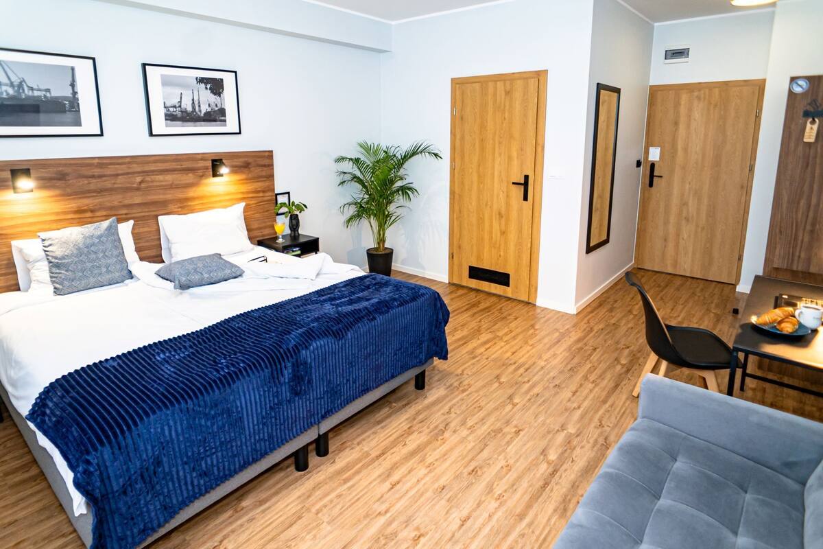 Et moderne hotelværelse med trægulv, en seng med et blåt tæppe, et skrivebord med stol og en potteplante.