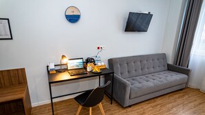A modern living room with a grey sofa, a wooden floor, a desk with a laptop, a chair, a clock, and a TV mounted on the wall.